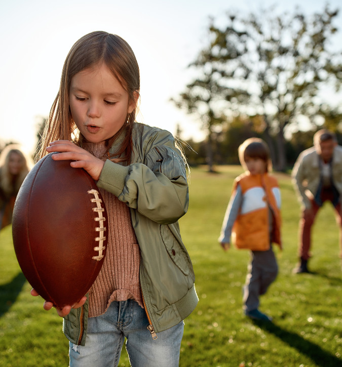 young girl holding football on its tips with family ready to play ball behind her