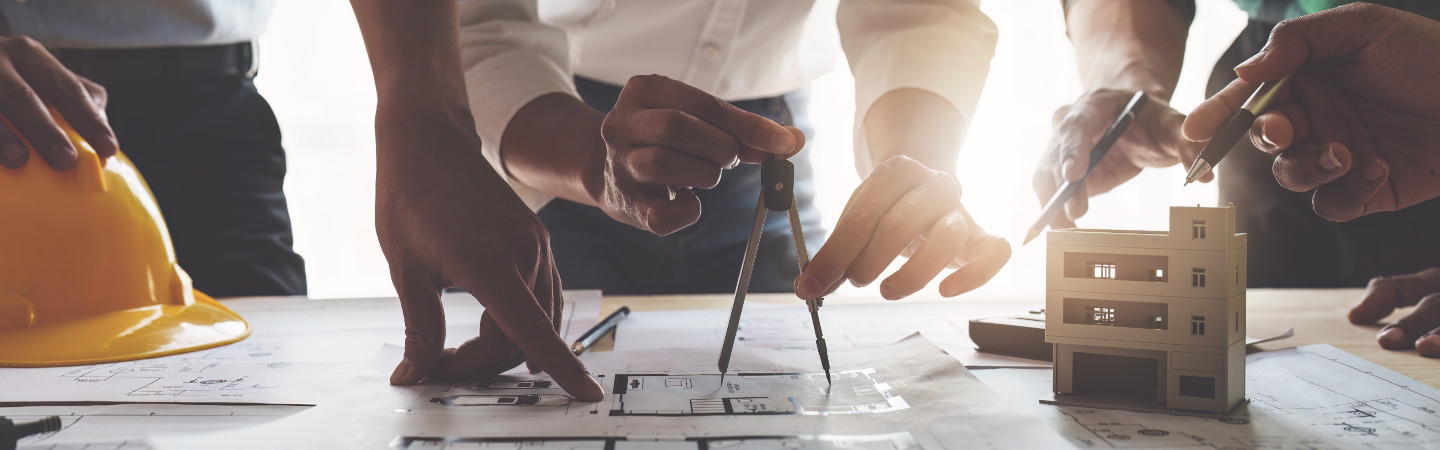 Several sets of hands around a drafting table, one person is holding a drafting tool, another is holding a hard hat; small replica of building - all sitting on plan blueprints