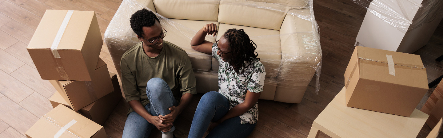 African American couple sitting on the floor amid moving boxes.  Both are smiling and looking at each other.