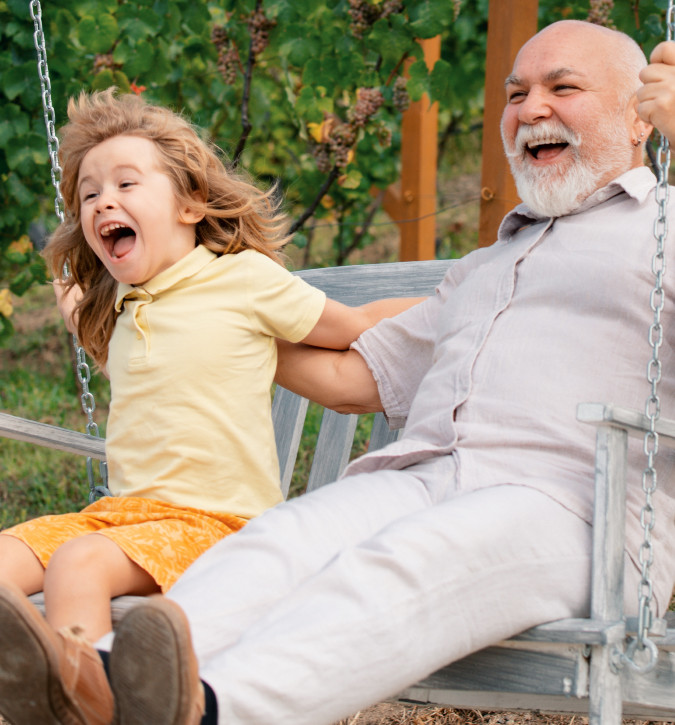 Older gentleman swinging on wooden swing with young child.  Both are smiling and laughing.