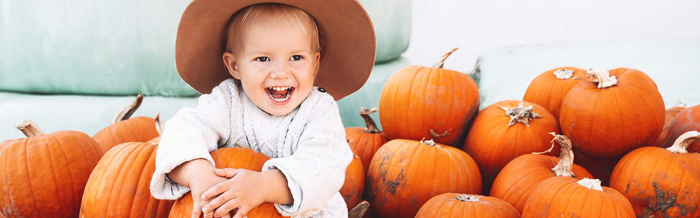 young child wearing floppy hat, siting holding a pumpkin with pumpkins all around him