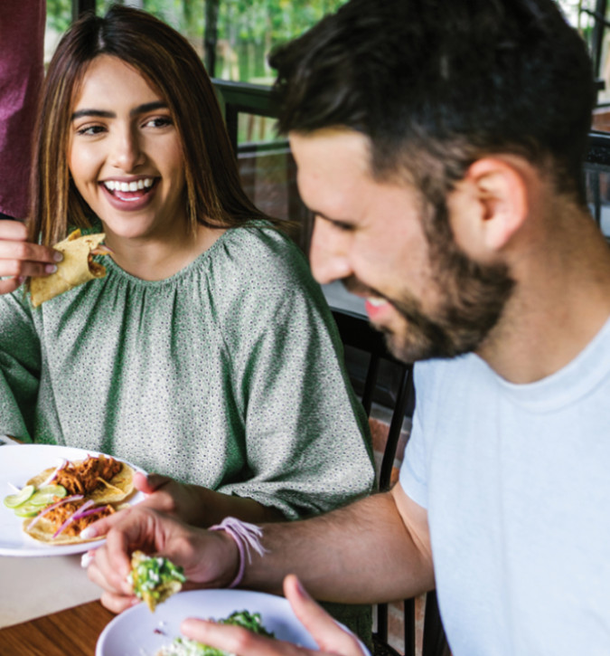 Lady smiling while eating a taco with a man.  Both are eating tacos and sitting at a table.