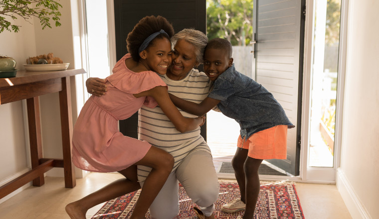 Grandmother kneeling in entry way hugging both of her grandkids; all are smiling and happy