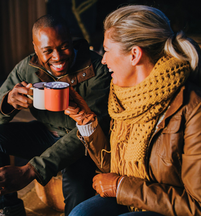 Two adults, dressed in coats, sitting near campfire, toasting with coffee mugs