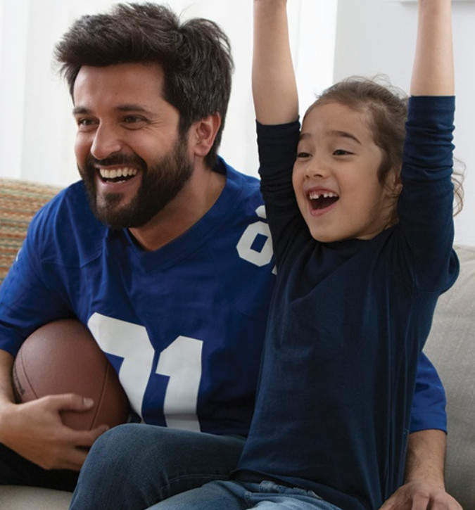Man and young girl, both smiling, sitting on couch cheering.  Man is holding a football.  Young girl is cheering with both arms raised.