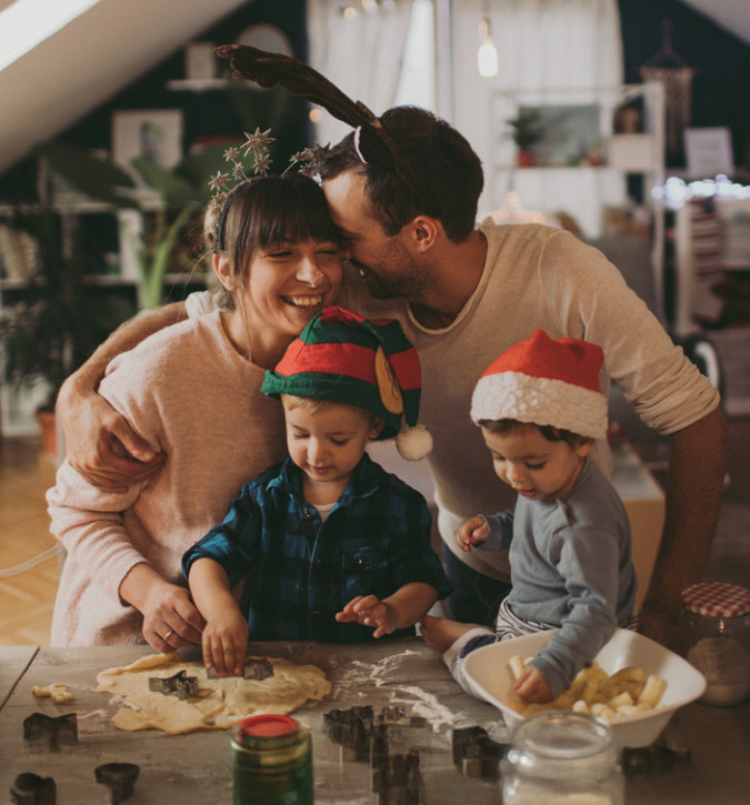 Man and lady standing behind two small children.  All standing at counter rolling cookie dough and  using cookie cutters.  Smiling and wearing Christmas hats.