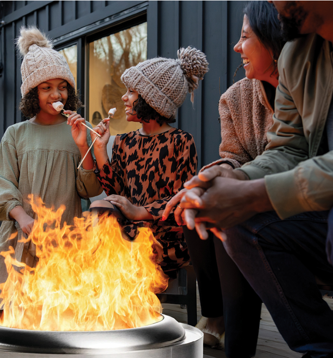 Two young girls wearing winter caps, both are giggling and eating roasted marshmallows.  They are seated around fire with two adults who are also smiling.  