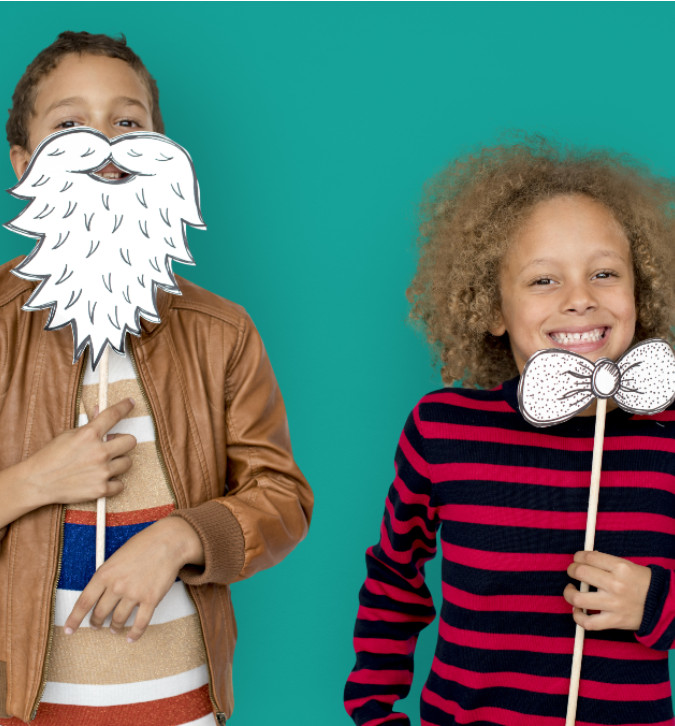 Two young children posing with photo props. One child is holding a beard on a stick up over his lower face. The other child is holding a bowtie.