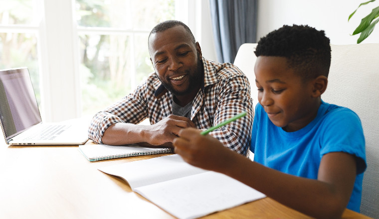 Dad smiling and helping young boy with is homework, both are sitting at the table