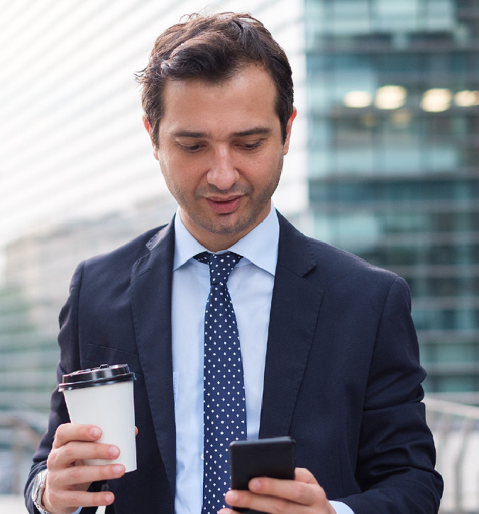 young professional man wearing a suit, holding cup of coffee, looking at his cellphone