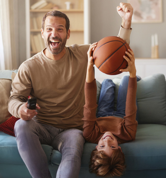 Man is sitting on the couch next to his son.  The man is holding the TV remote in one hand and raising a fist pump with the other arm.  The young son is laying upside down on the couch and is holding a basketball.  Both are smiling and may be cheering on their favorite team that is playing on the TV.