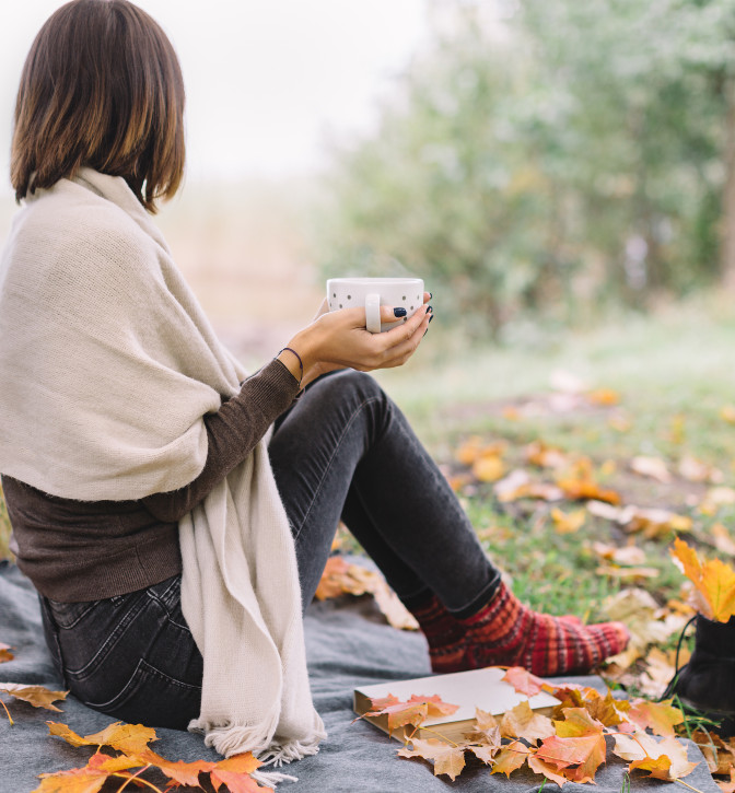 Lady holding a cup of coffee, wearing a shawl, looking away from the camera.  She is sitting outside on a blanket with her shoes off.  Fall leaves are all around her.