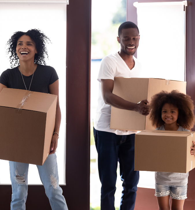Family of three walking through front door, all holding moving boxes and smiling