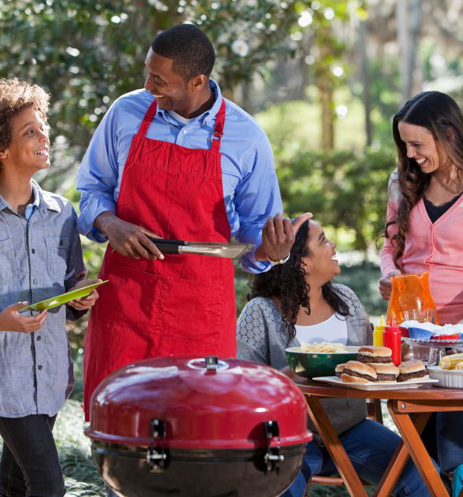 Family gathered around outdoor grill.  Man in red apron smiling; boy holding plate smiling.  Young lady sitting at table with the food.  Lady smiling holding pitcher.