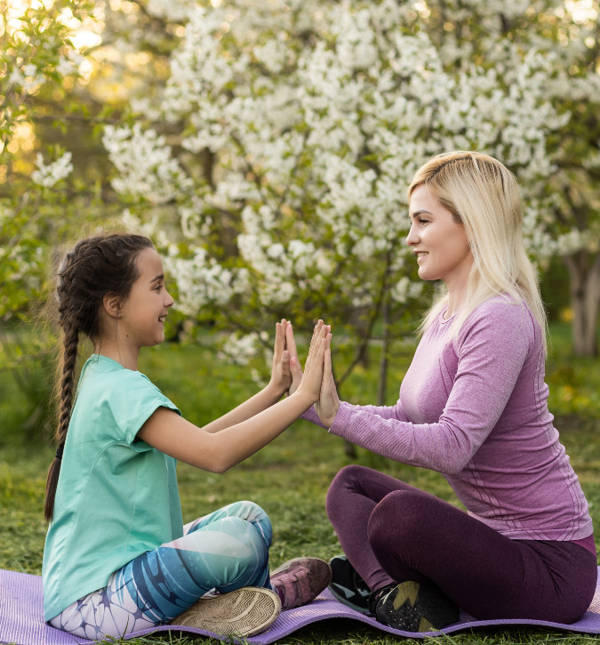 Mom and daughter sitting on exercise mat, both are sitting with crossed legs and facing each other, have their hands together, spring trees are blooming in background