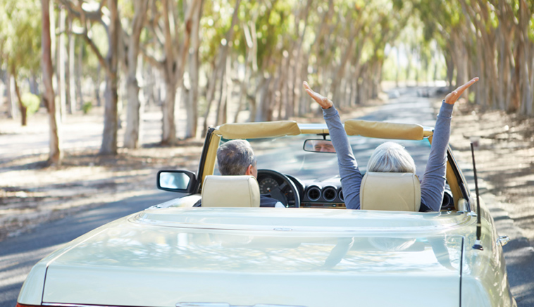 Retired couple driving away in convertible with top down.  Driving down lane of trees.